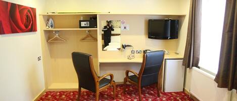 A hotel room with a red patterned carpet, two wooden chairs, a desk with a flat-screen TV, a small kitchenette, and a large window with curtains.