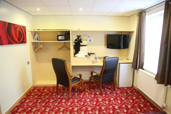 A hotel room with a red patterned carpet, two wooden chairs, a desk with a flat-screen TV, a small kitchenette, and a large window with curtains. - The Cambridge Hotel (Huddersfield)