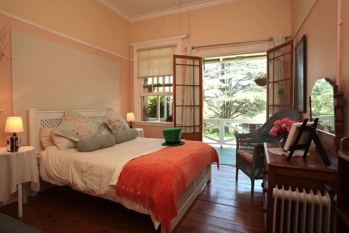 Un dormitorio con cama, mesitas de noche, una silla, un radiador y una ventana con vistas al verdor.