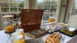 A breakfast buffet with various food items, including pastries, fruits, and beverages, set up in a room with large windows. - Peterstone Court (Brecon)