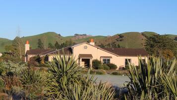 A two-story house with a front porch, surrounded by lush greenery and hills in the background.