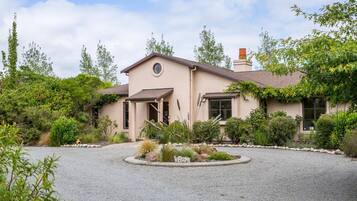 A single-story house with a circular driveway, surrounded by various shrubs and trees.
