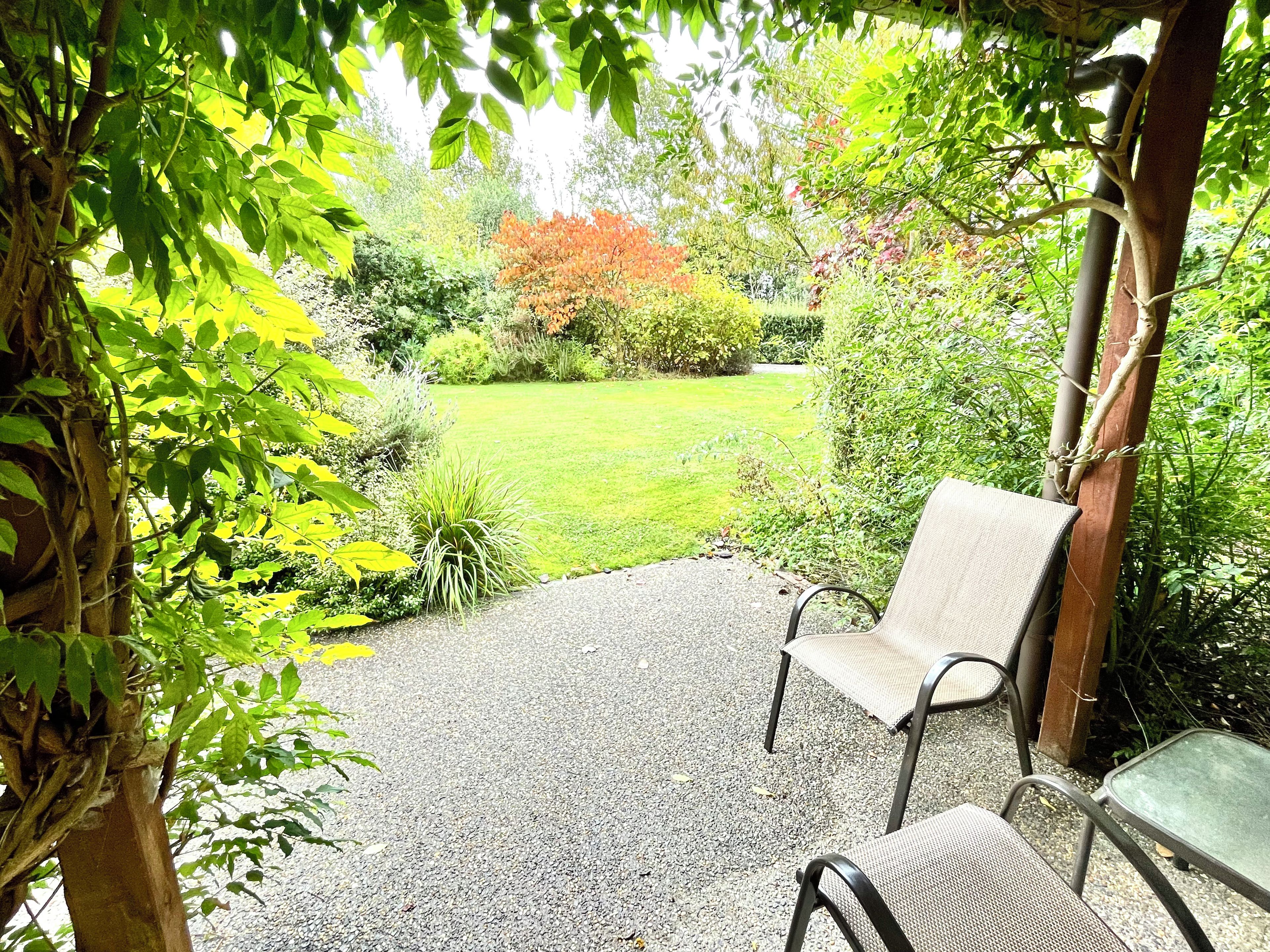 A covered patio area with a gravel surface, two metal chairs, and a view of a grassy garden with various plants and a tree.