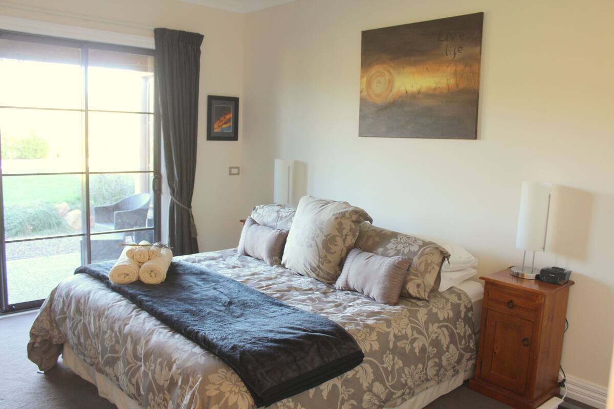 Quarto com cama, mesa de cabeceira, janela com cortinas e um quadro na parede.