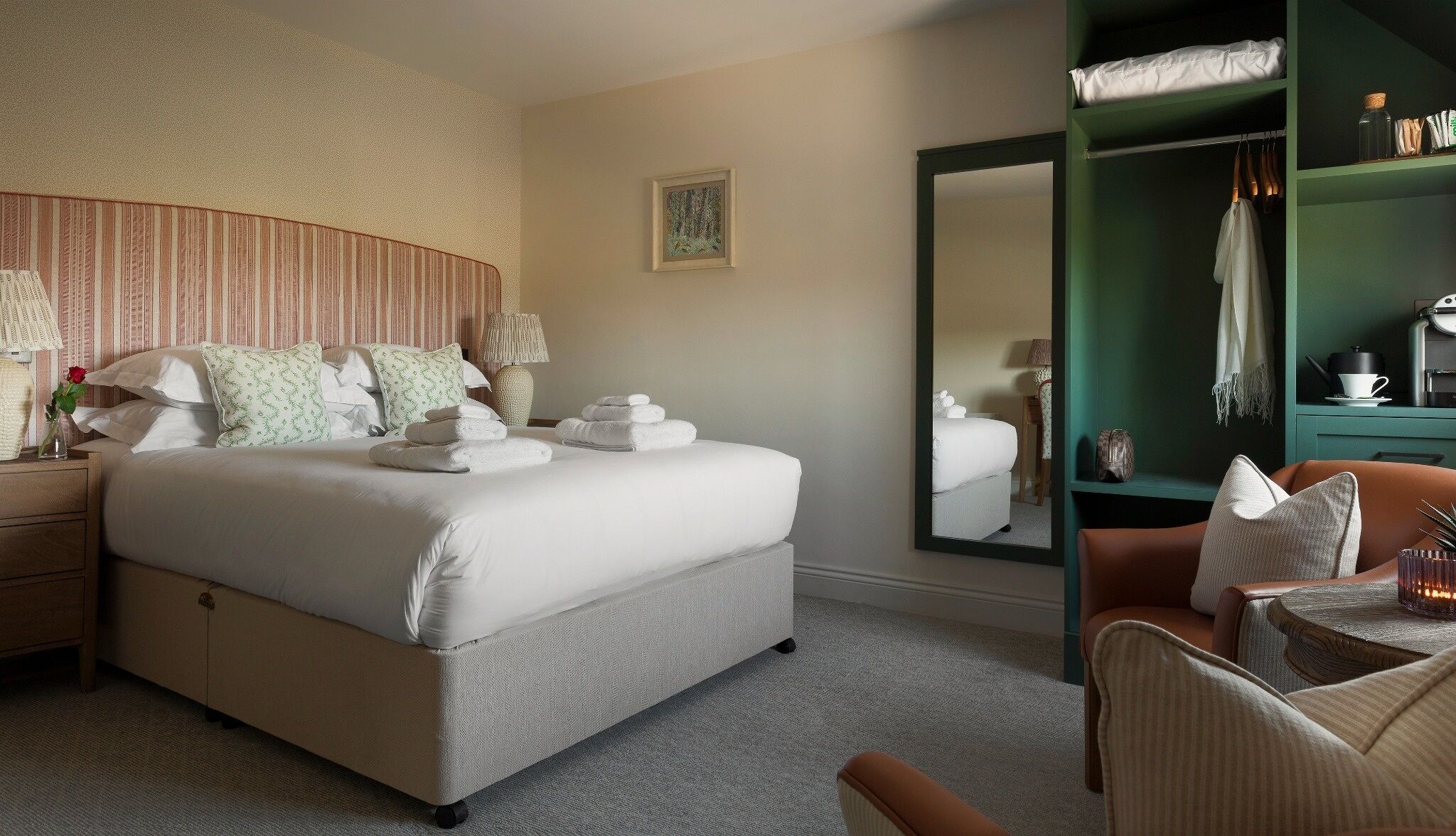 Una cama bien hecha con ropa blanca y una pila de toallas dobladas, una mesita de noche con una lámpara y un florero con una sola rosa, un sillón tapizado en verde y un armario verde con espejo.