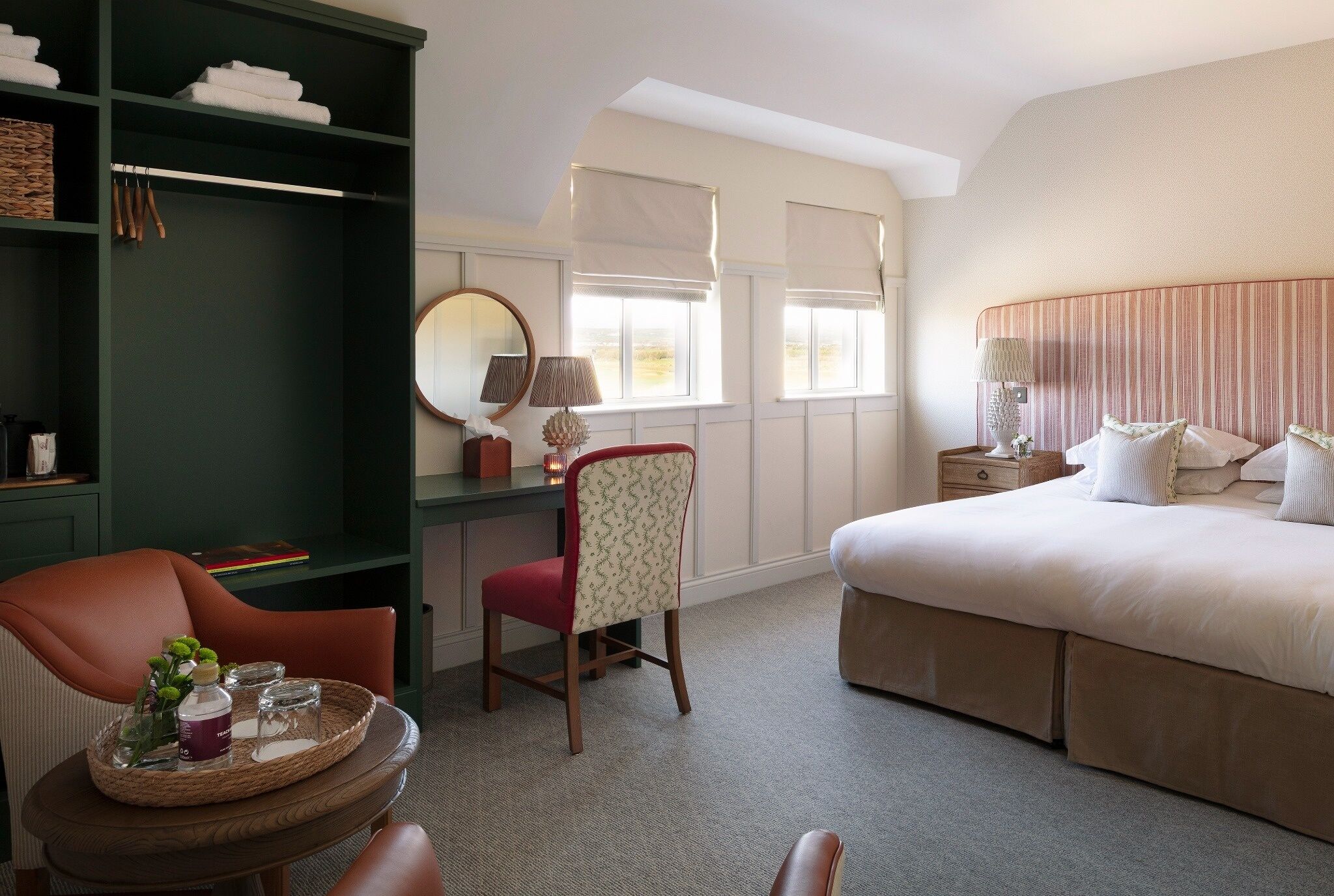 A hotel room with a bed, a desk with a chair, a round mirror, and a small table with a tray.
