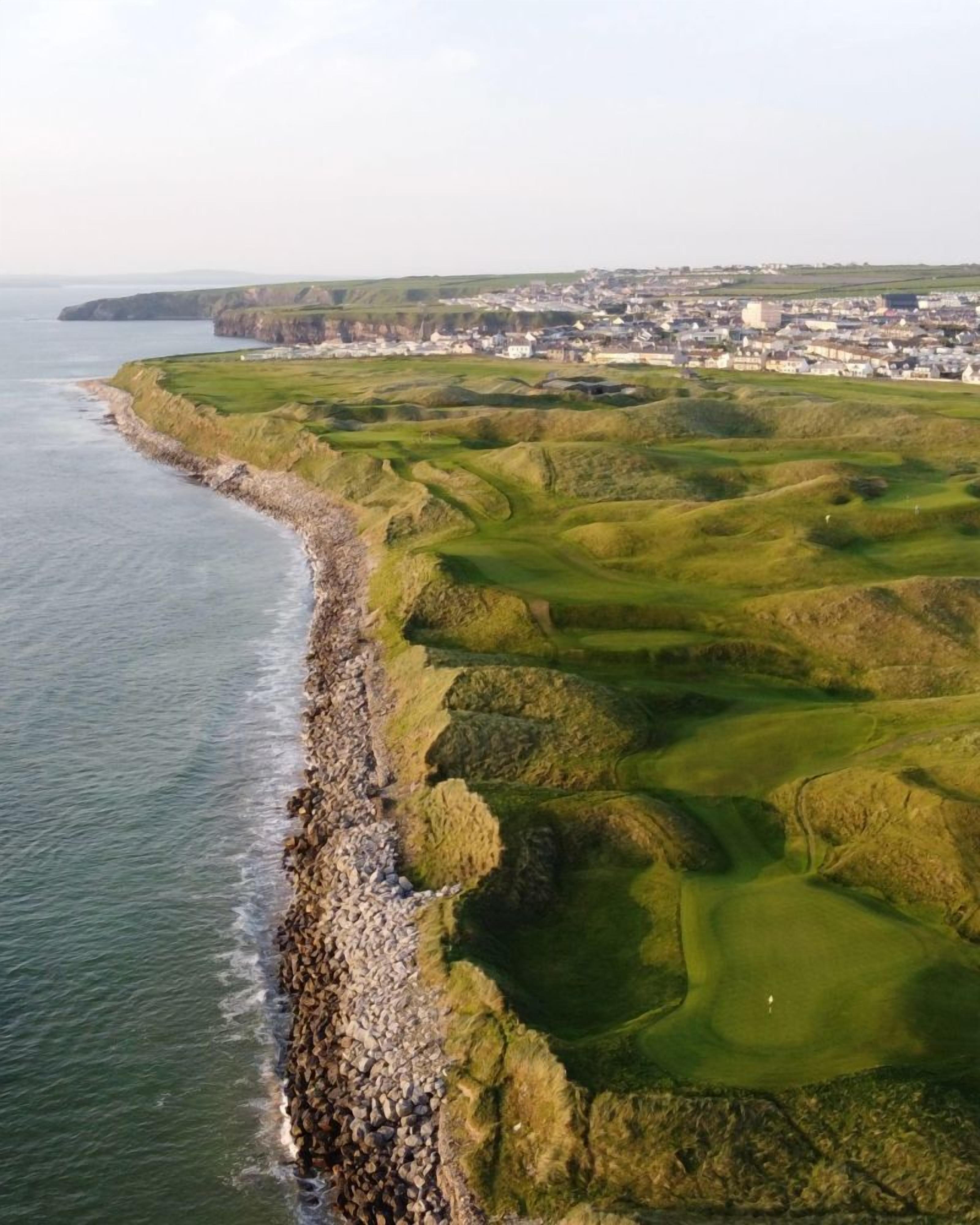 An aerial view of a coastal golf course with a rocky shoreline and a town in the background.