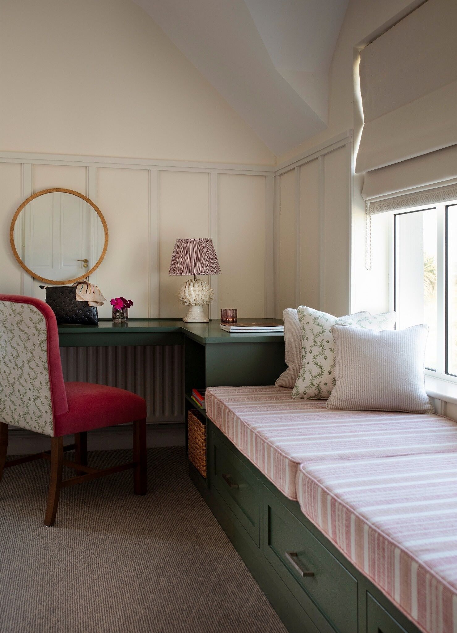 A bedroom with a green dresser, a red chair, a bed with striped bedding, a round mirror, and a lamp.