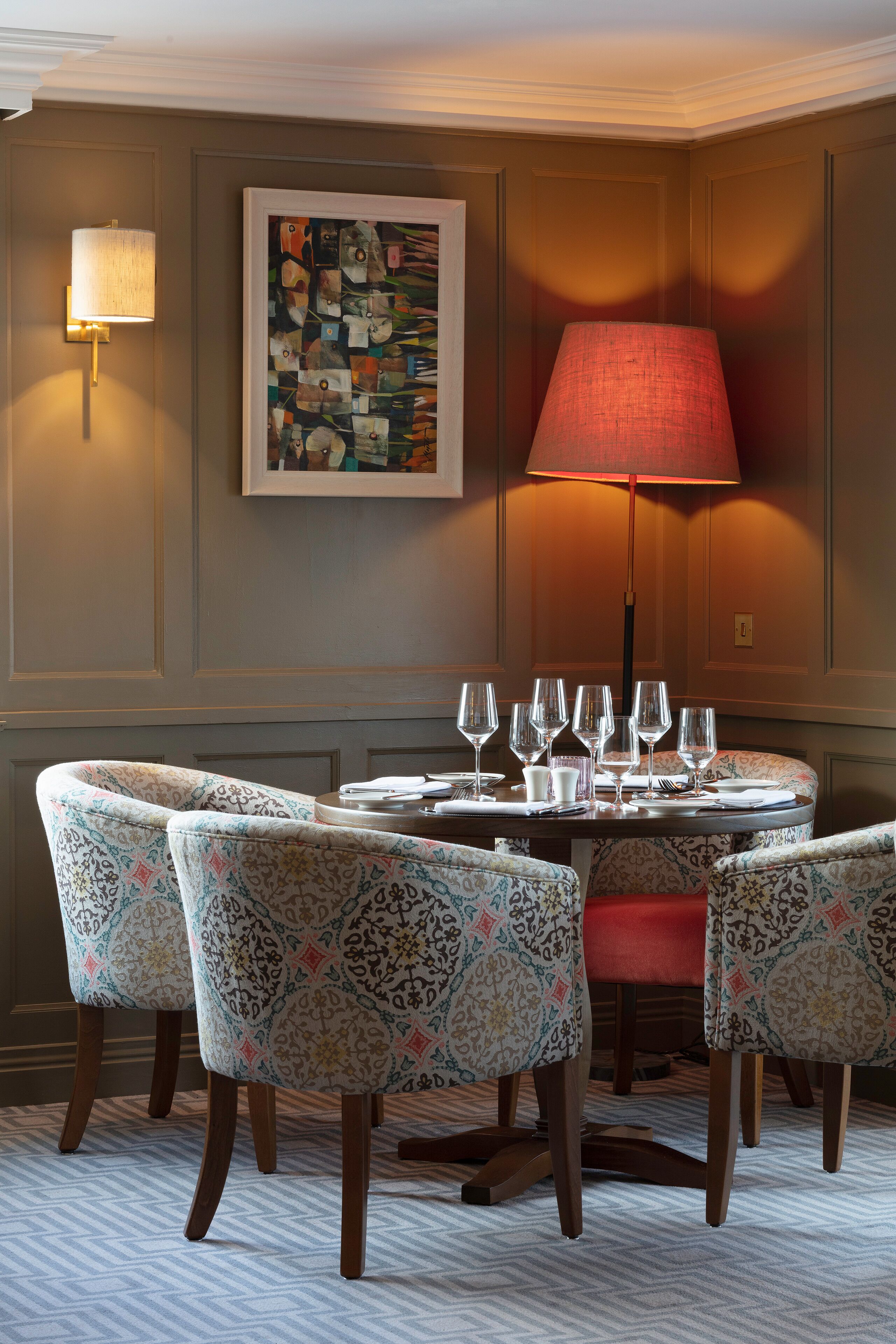 A dining area with a round table, six patterned chairs, a red lamp, and a framed abstract painting on the wall.