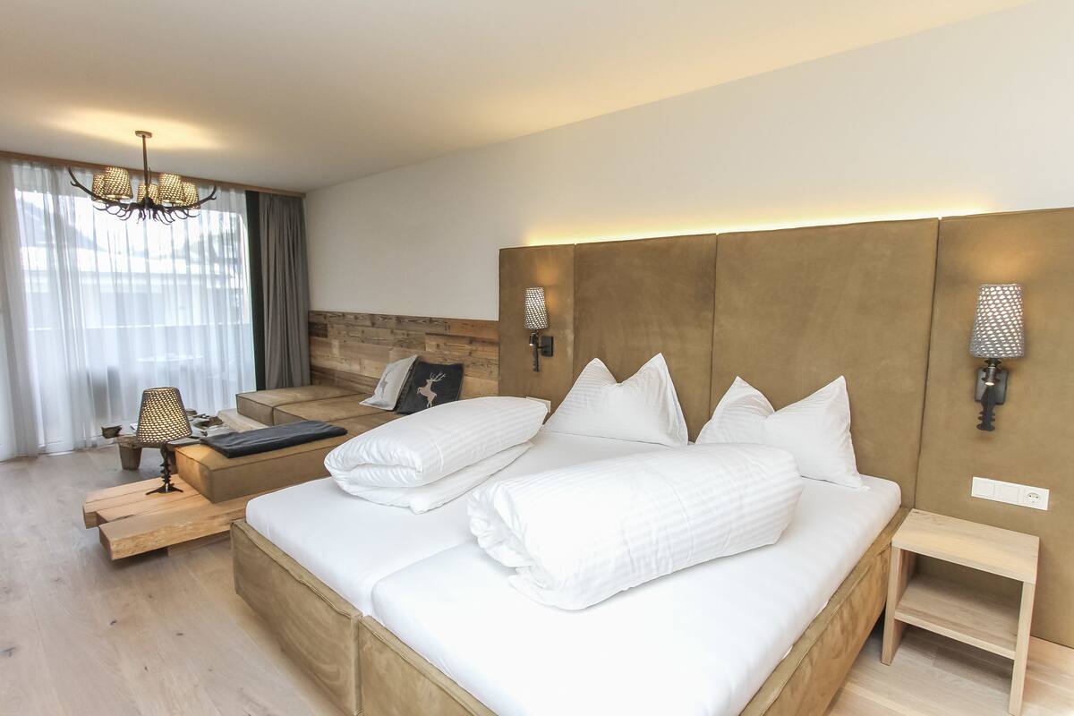 Une chambre d’hôtel moderne avec un grand lit, une tête de lit en bois, une table de chevet, une lampe et une vue sur l’extérieur à travers des rideaux transparents.