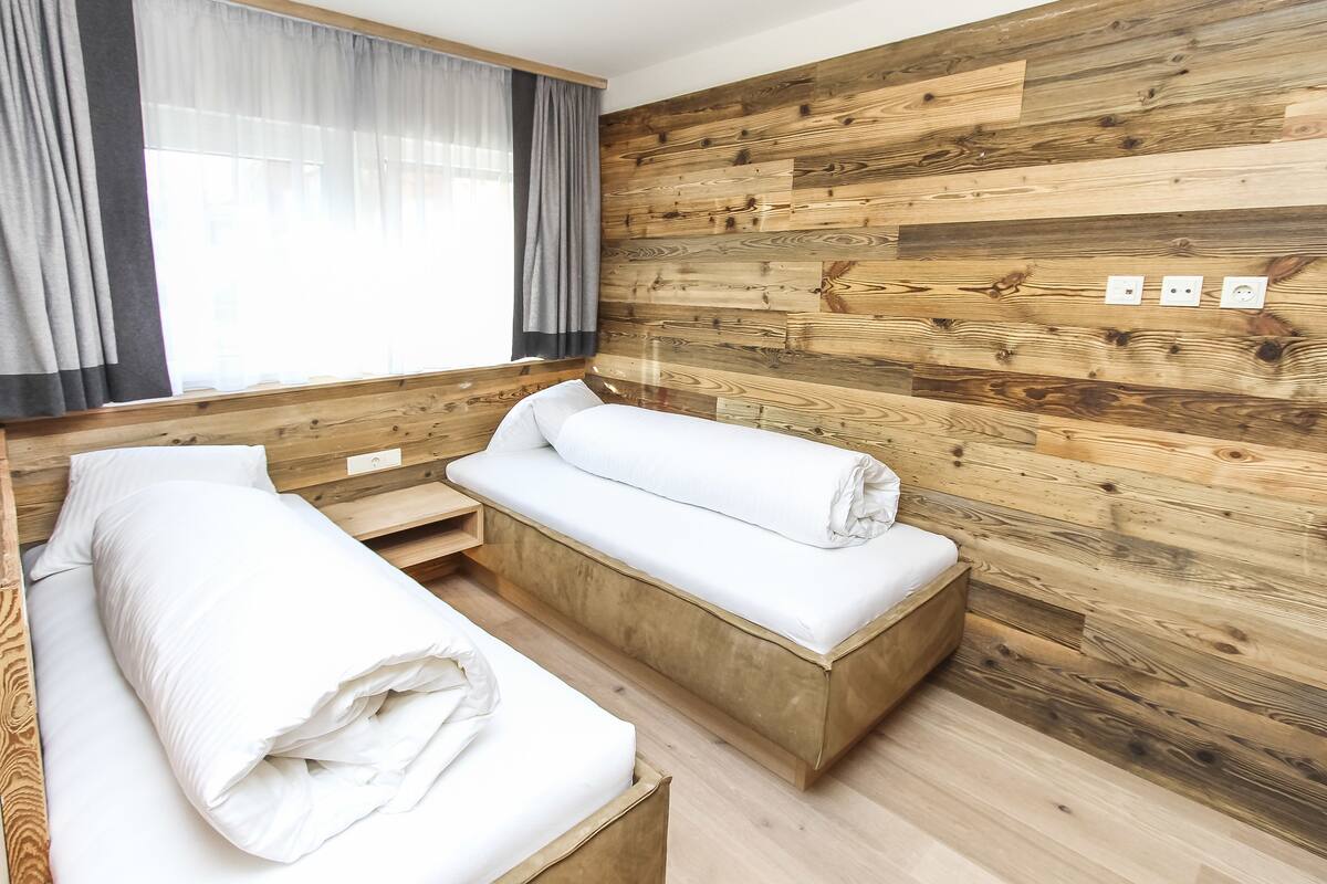 Une pièce avec deux lits en bois, une fenêtre avec des rideaux légers et un mur en bois équipé de prises électriques.