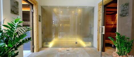 A modern hotel lobby with a glass-enclosed elevator, wooden flooring, and potted plants.