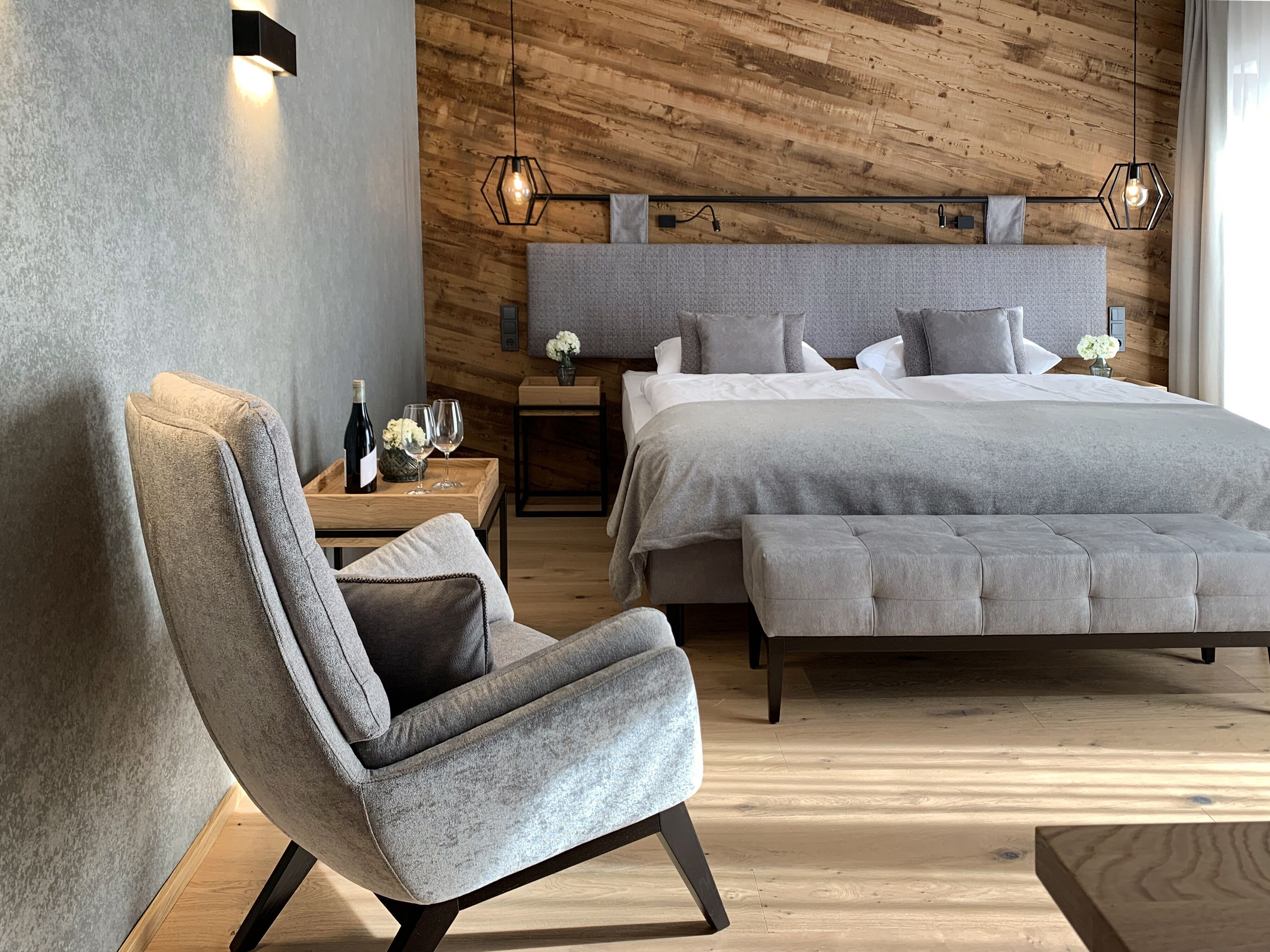 A modern bedroom with a wooden headboard, a grey upholstered chair, a large bed with multiple pillows, and a bedside table with a bottle and glasses.