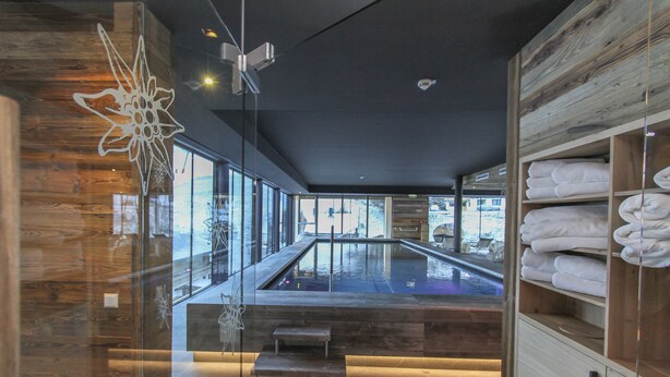 Un área de spa moderna con un hidromasaje envuelto en cristal, estanterías de madera con toallas y vistas a la ciudad.