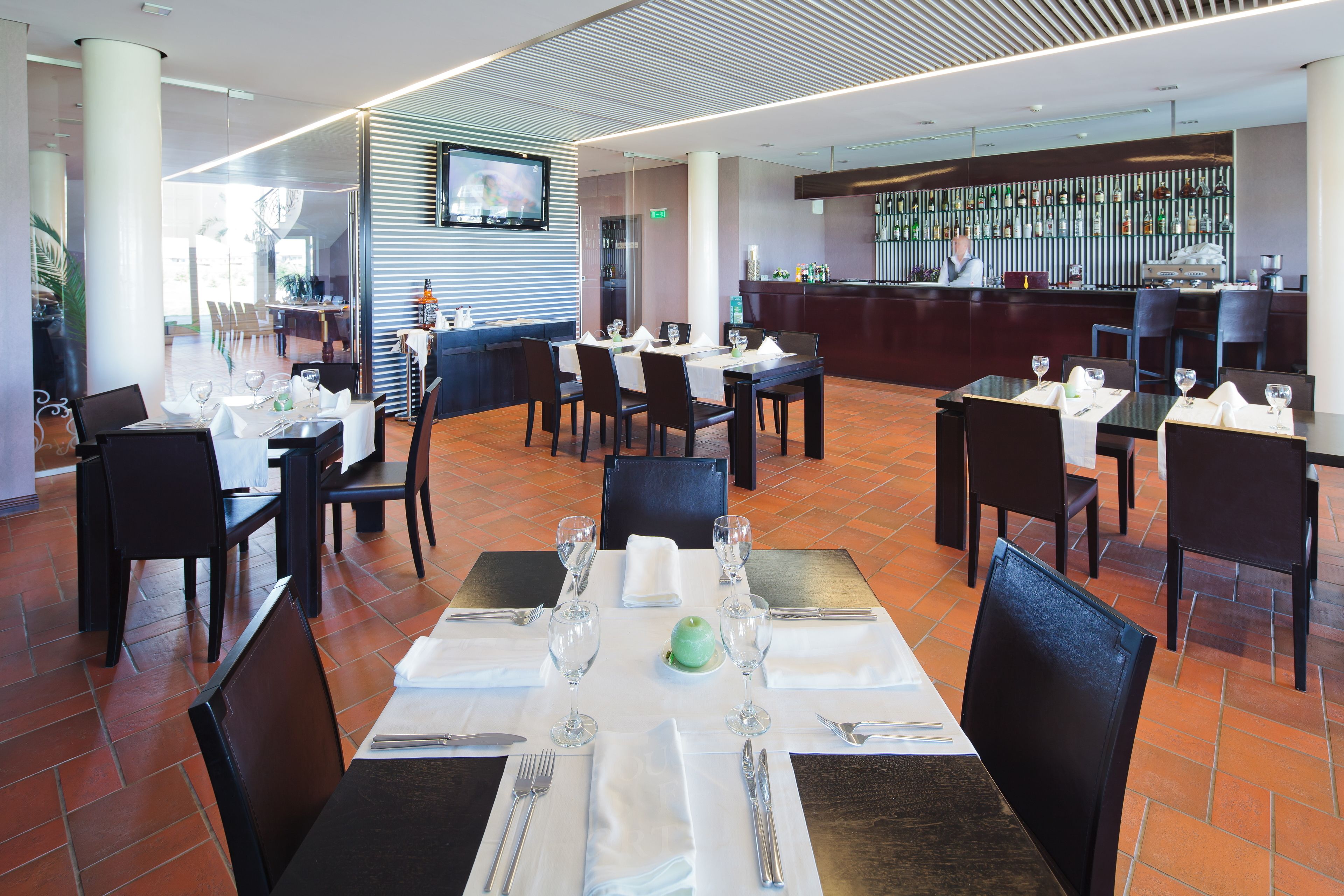 A modern restaurant with multiple dining tables set for guests, a bar area, and a television screen mounted on the wall.