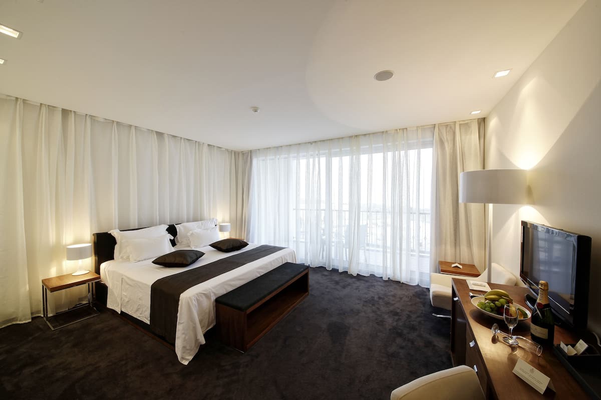 A hotel room with a large bed, a desk with a television, a minibar, and a view of the city through large windows.