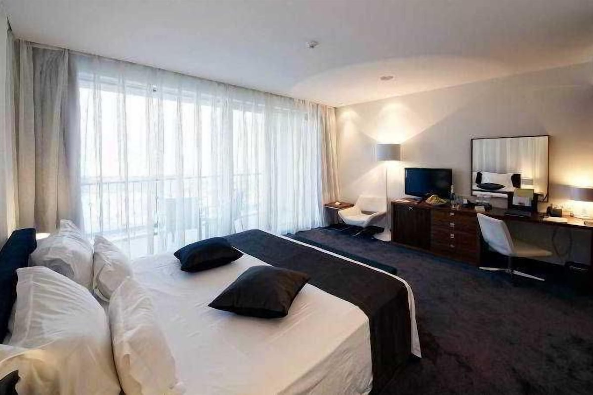Habitación de hotel con cama, escritorio, silla, televisión y un ventanal con cortinas.