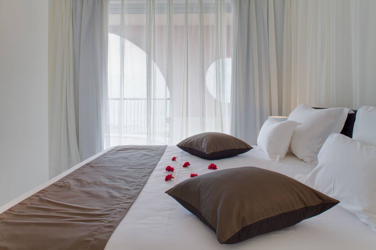 A neatly made bed with white and brown bedding, a window with sheer curtains, and a view of the outside.