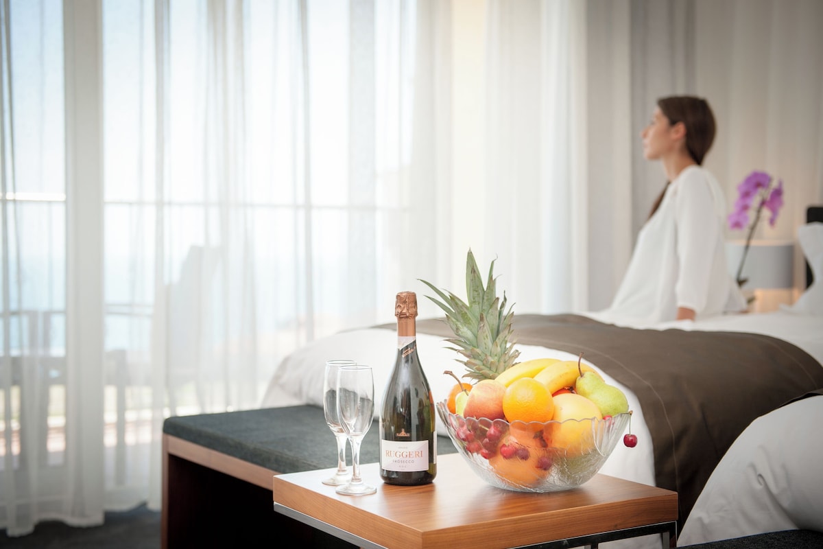 Una habitación de hotel con una cama, una mesita de noche con una botella de vino, copas y un frutero.