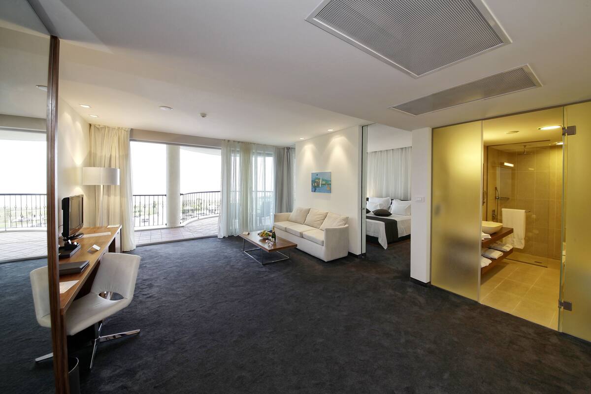 A modern hotel room with a dark carpet, a white sofa, a desk with a chair, a TV, and a large window with curtains.
