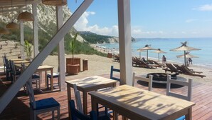 Un restaurante frente a la playa con terraza, vistas a la orilla y montañas al fondo.