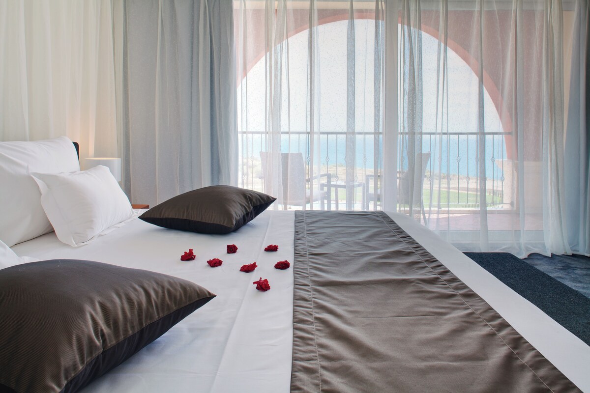 Habitación de hotel con cama, ropa de cama blanca y marrón, y vistas al mar a través de amplias ventanas.