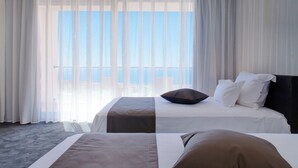 Habitación de hotel con una cama grande, una cama más pequeña y vistas al mar a través de cortinas translúcidas.