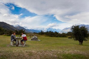 Una mesa de picnic con dos personas sentadas, rodeada por un campo de césped y montañas al fondo.