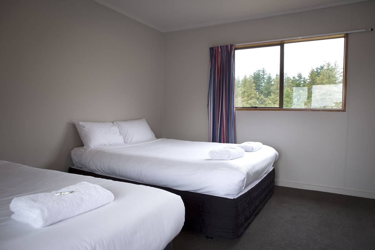 A hotel room with two beds, a large window with curtains, and a view of trees outside.