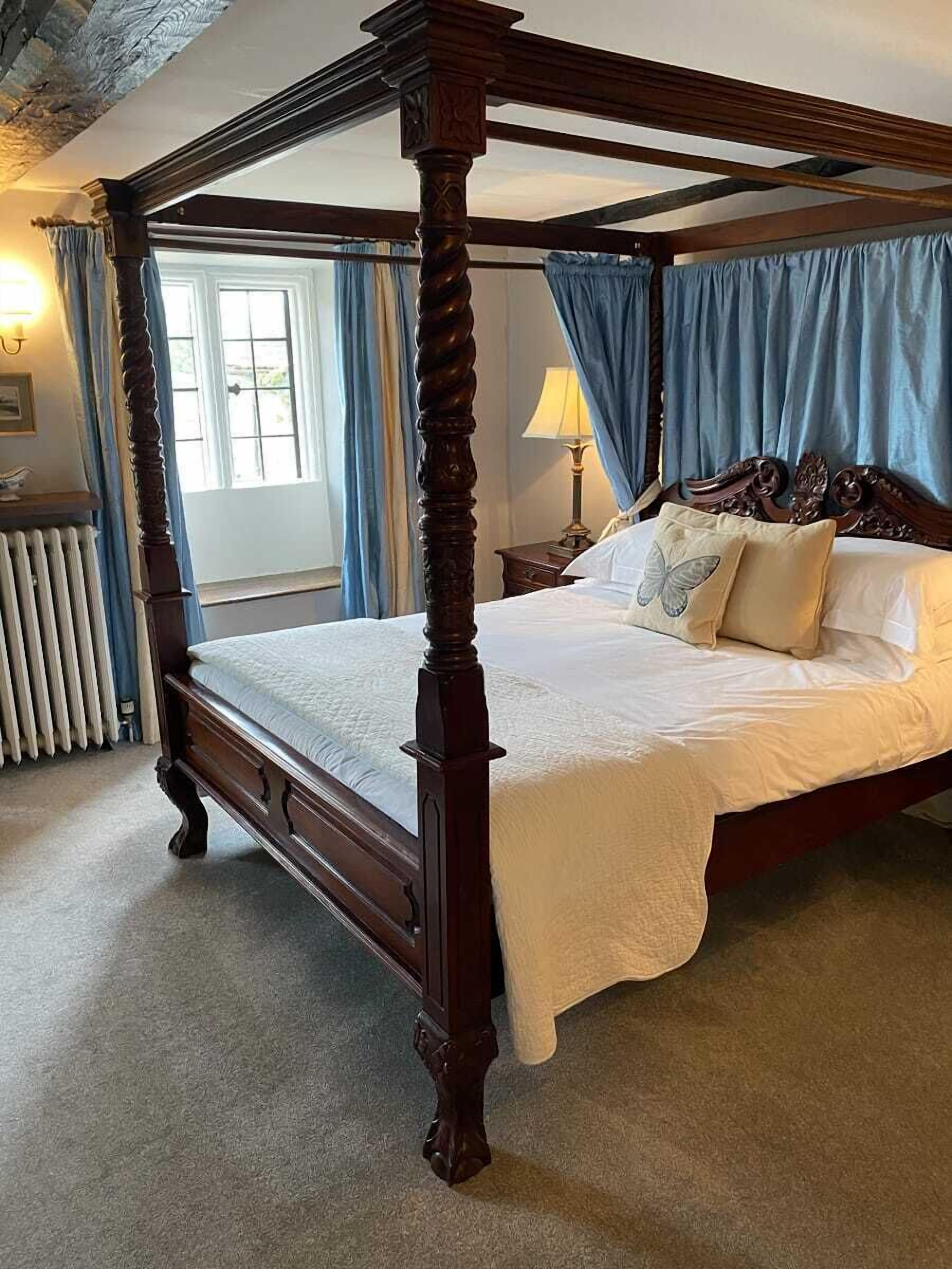 A four-poster bed with a wooden frame and white bedding, a radiator, and a window with curtains.