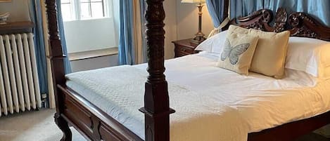 A four-poster bed with a wooden frame and white bedding, a radiator, and a window with curtains.