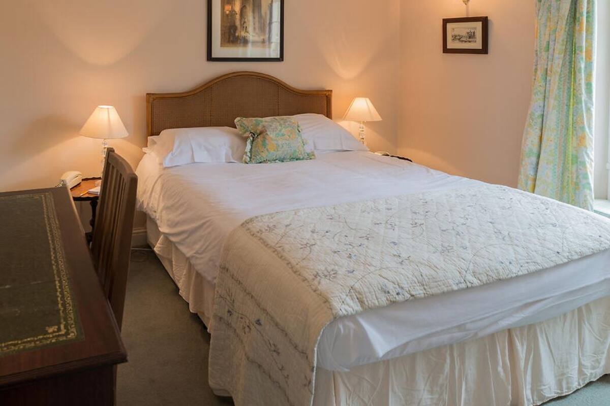 Una habitación de hotel con una cama grande, dos lámparas de noche, una mesita de noche de madera, un escritorio y un cuadro en la pared.