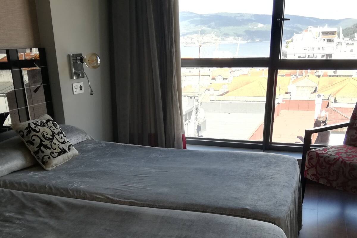 A hotel room with two beds, a chair, a large window with a view of buildings and hills, and a wall-mounted lamp.