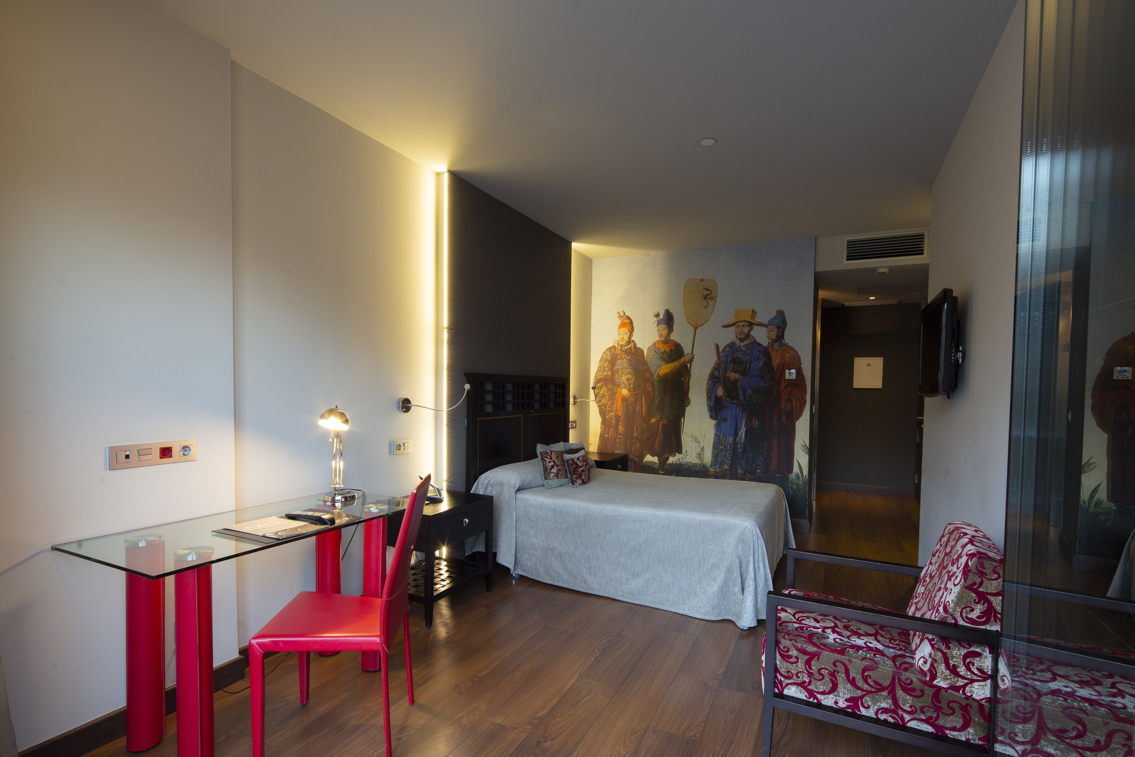 Una habitación de hotel moderna con un escritorio de vidrio, una silla roja, una cama con manta estampada y un mural en la pared.