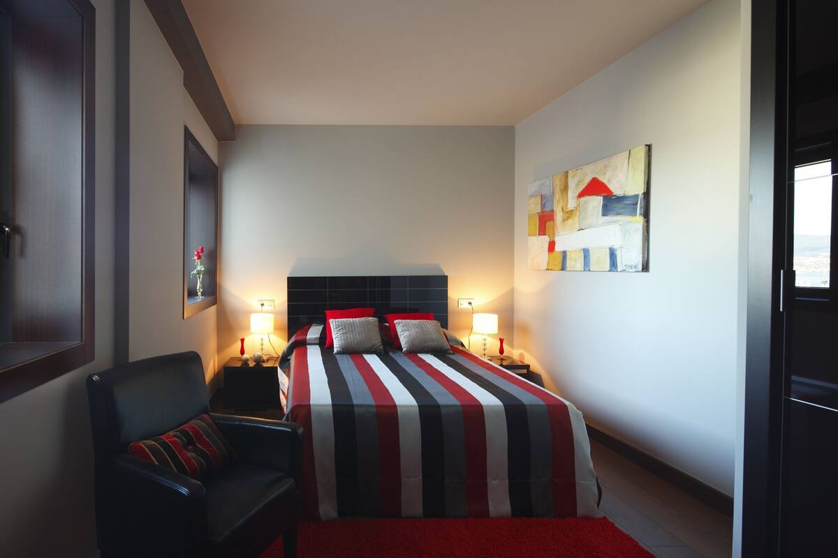 A hotel room with a bed, a chair, a nightstand, a lamp, and a painting on the wall.