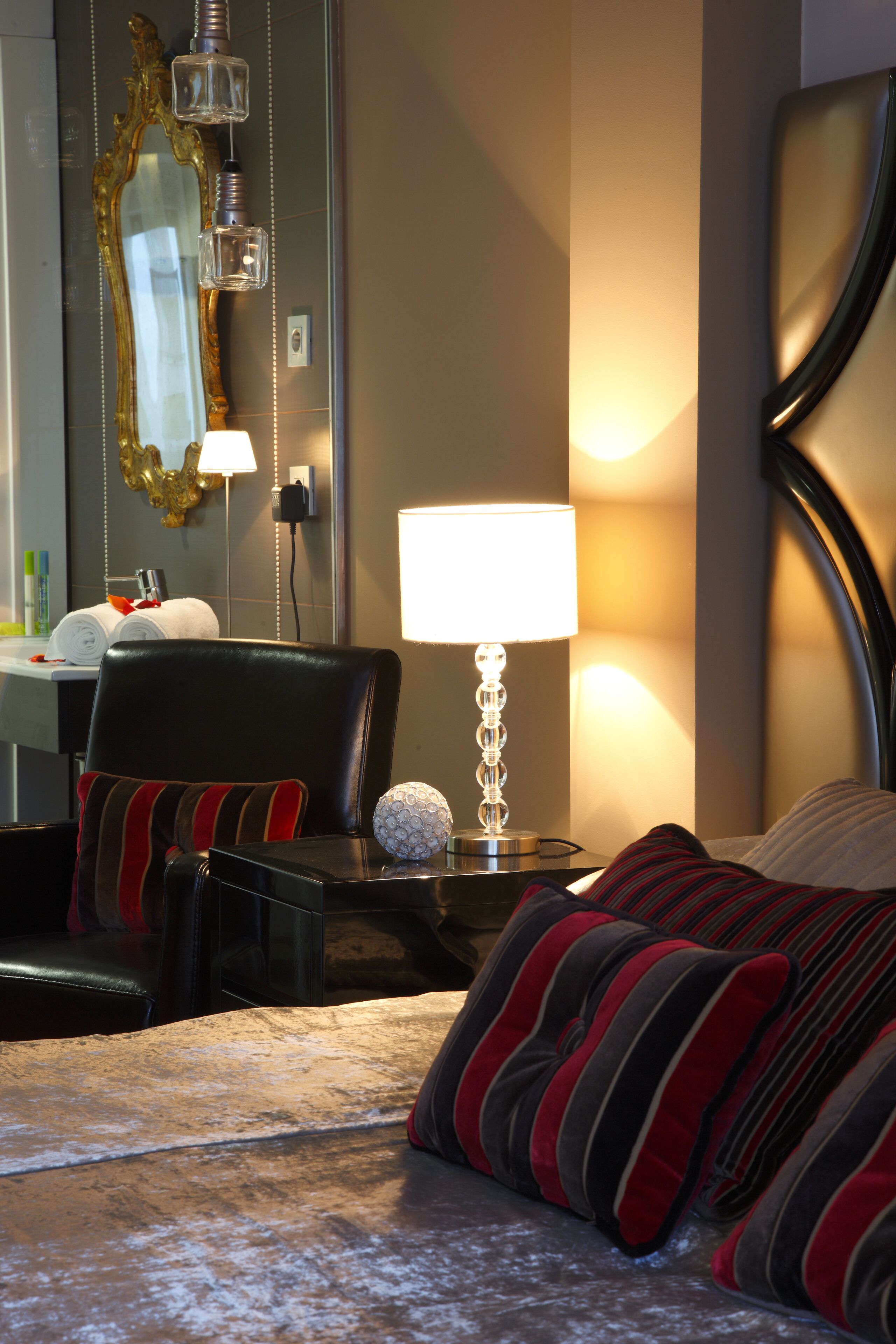 A hotel room with a bed, a chair, a nightstand with a lamp, a mirror, and a wall-mounted light fixture.