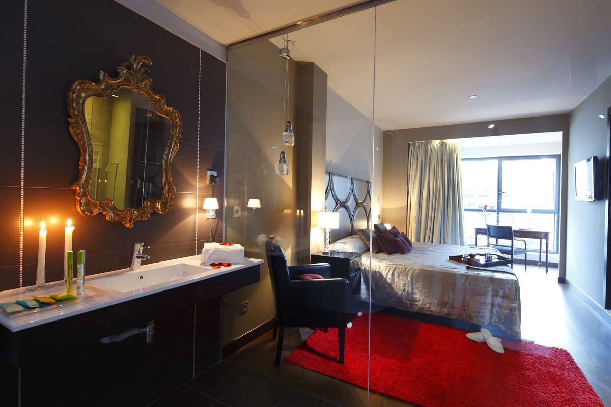 A modern hotel room with a large mirror, a bed with a red rug, a desk, and a view of the outdoors.