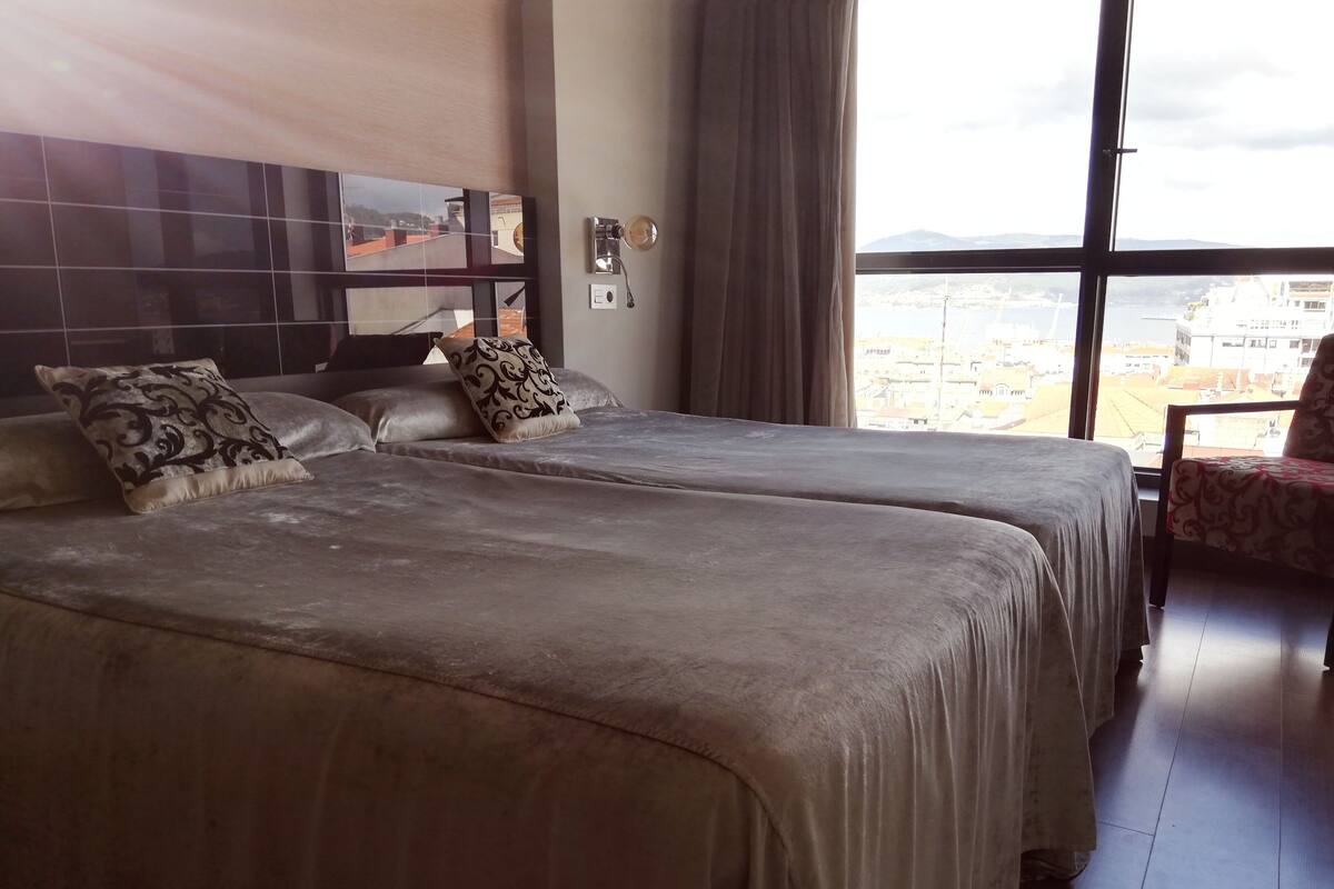 A hotel room with two beds, a large window with a city view, and a chair with a patterned cover.