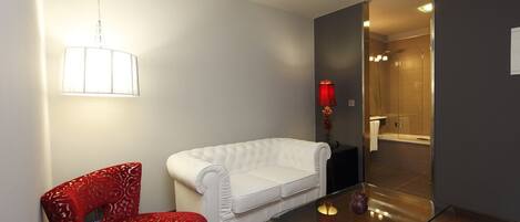 A modern room with a white sofa, a red patterned chair, a glass table, and a shower area visible through a glass door.
