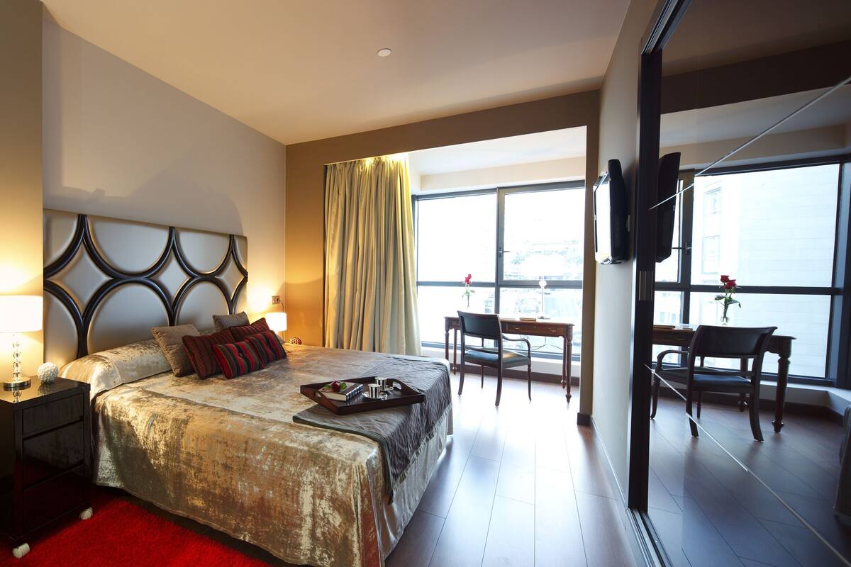 A hotel room with a bed, bedside tables, a desk, and a chair. There is a large window providing a view of the outside.