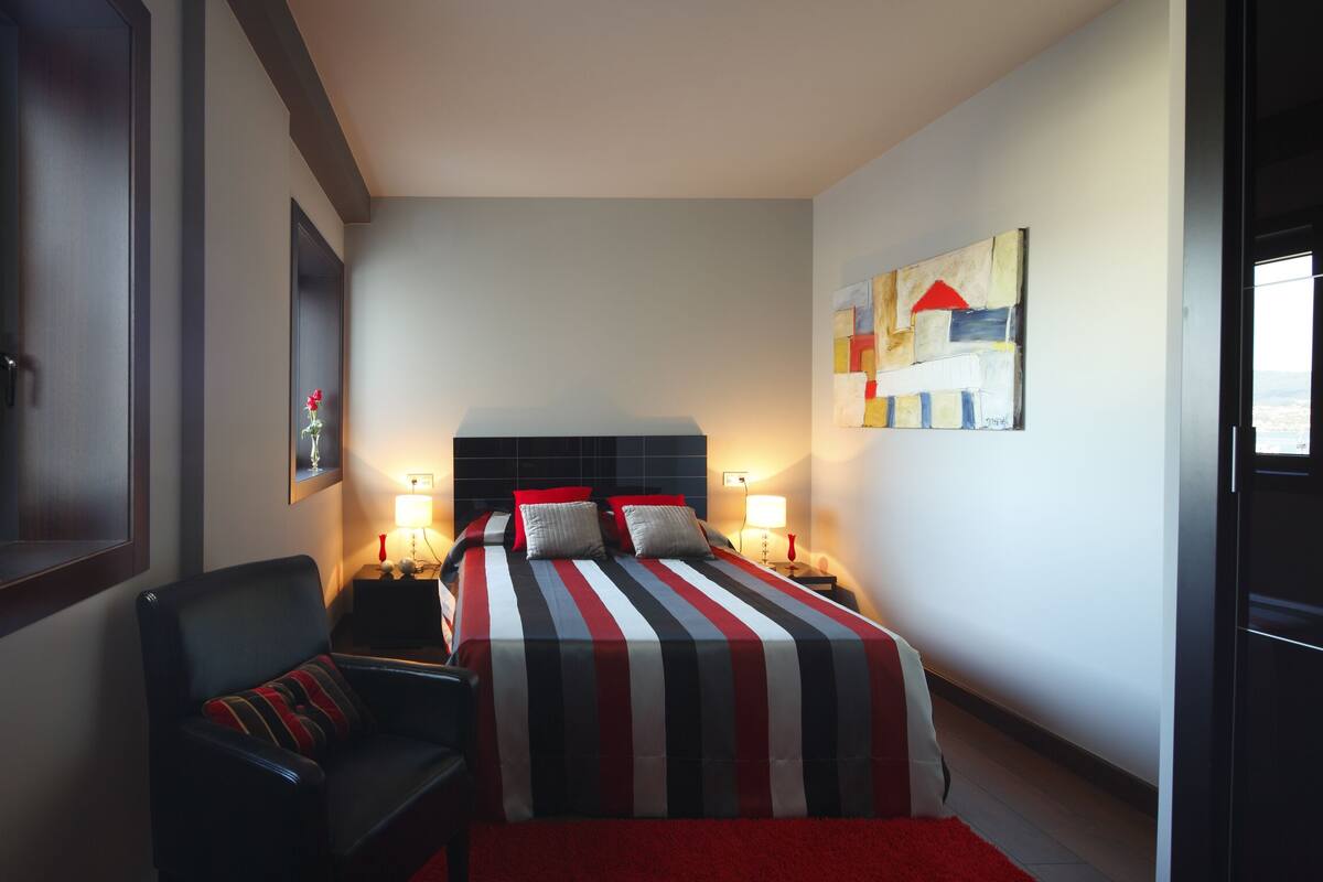 A hotel room with a bed, a chair, a nightstand, a lamp, and a painting on the wall.
