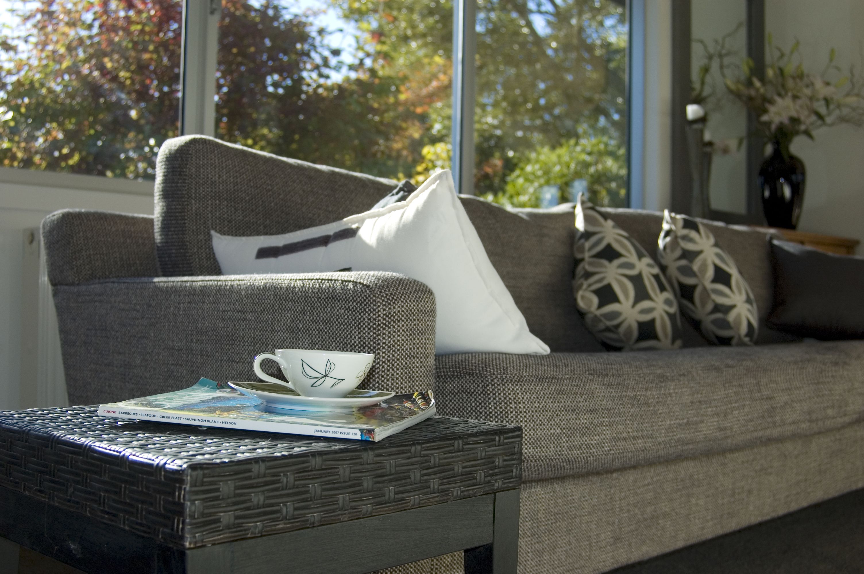 A cozy living area with a grey sofa, a wicker coffee table, and a window with a view of trees.