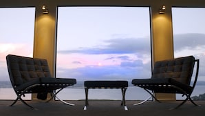 Una habitación con dos sillas negras y una mesa pequeña, ventanas amplias con vistas a la puesta de sol sobre el agua.