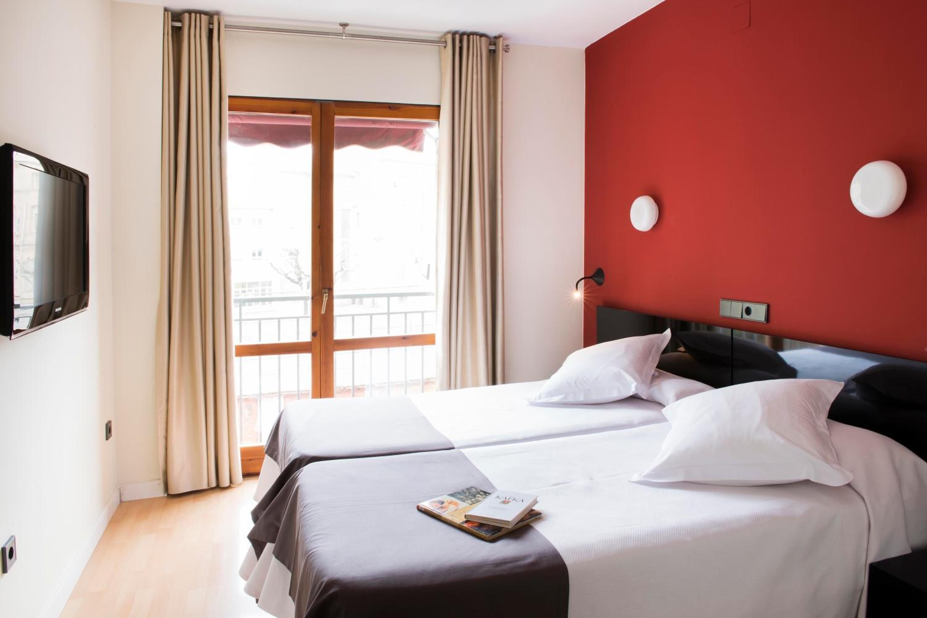Een hotelkamer met twee bedden, een televisie, een rode accentmuur en een balkon met gordijnen.