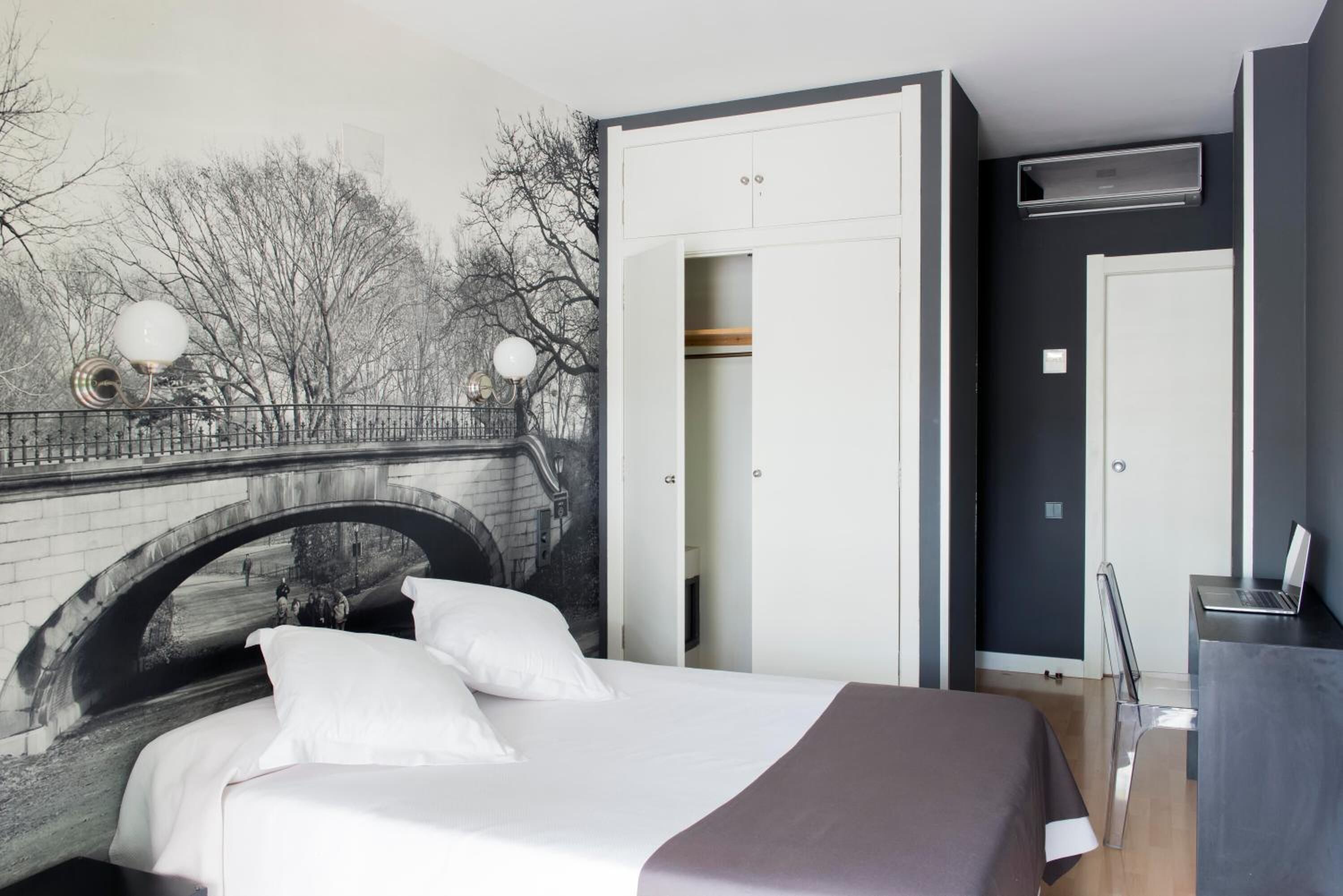Un dormitorio con una cama, un armario, un escritorio con una computadora portátil y un mural de un puente.