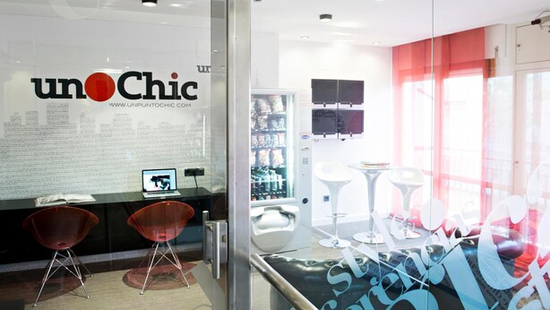 Un interior moderno con una pared de vidrio mostrando el logotipo 'unOChic' y una dirección web. Hay dos sillas rojas, un taburete blanco y una silla blanca cerca de una pared divisoria de vidrio.
