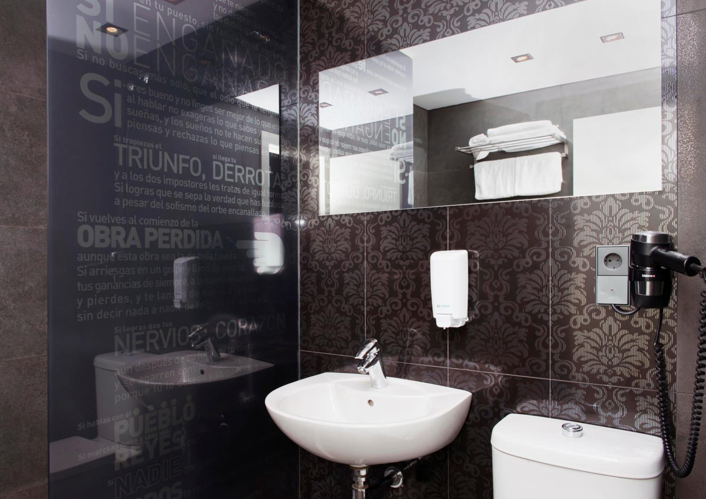 Un baño con lavamanos, inodoro y espejo. Hay un dispensador de desinfectante de manos y un secador de pelo en la pared.