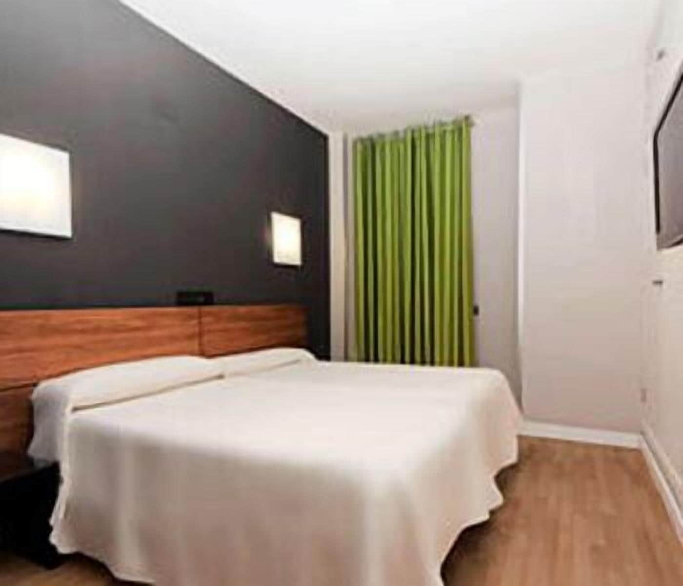 Ein Hotelzimmer mit einem Bett, einem Holz-Kopfteil, einem grünen Vorhang und einer weißen Wand mit einer Lampe.