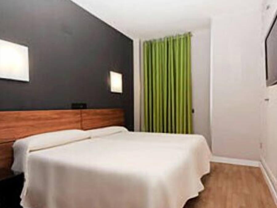 A hotel room with a bed, a wooden headboard, a green curtain, and a white wall with a light fixture.