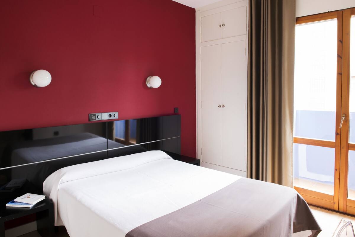 Une chambre d’hôtel comprenant un lit, une table de chevet, un miroir, une armoire et une fenêtre avec des rideaux.